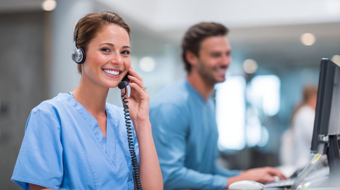 Smiling healthcare staff member in scrubs wearing a headset and speaking on the phone at a front desk workstation